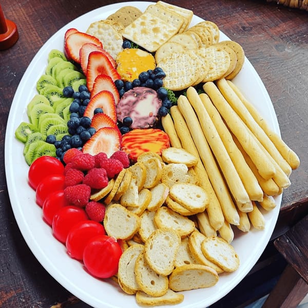 A colorful platter of sliced kiwi, strawberries, blueberries, raspberries, cheeses, crackers, and breadsticks.