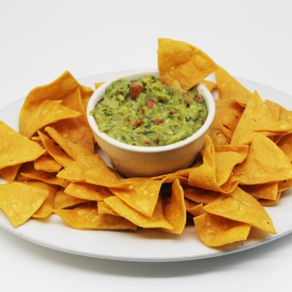 A white bowl of chunky guacamole surrounded by crispy tortilla chips on a white plate.