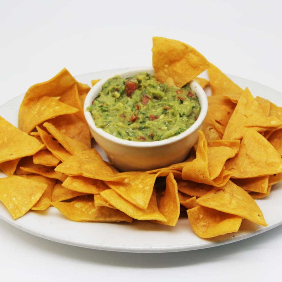 A white bowl of chunky guacamole surrounded by crispy tortilla chips on a white plate.